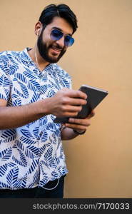 Portrait of young latin man using his digital tablet with earphones against yellow wall. Technology and urban concept.