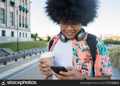 Portrait of young latin man using his mobile phone and holding a cup of coffee while walking outdoors on the street. Urban concept.
