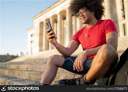 Portrait of young latin man using his mobile phone and wearing skate rollers while sitting outdoors. Sports concept. Urban concept.