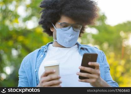 Portrait of young latin man wearing face mask and using his mobile phone while standing outdoors on the street. Urban concept.