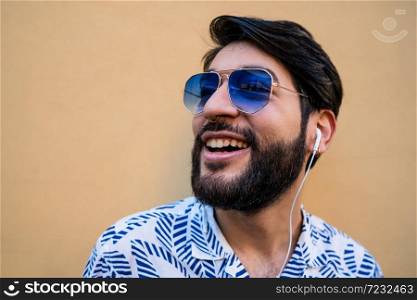 Portrait of young latin man wearing summer clothes and listening to music with earphones against yellow background.