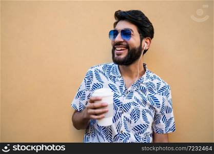 Portrait of young latin man wearing summer clothes, holding a cup of coffee and listening to music with earphones against yellow background.