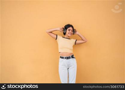 Portrait of young latin woman listening to music with headphones against yellow background. Urban concept.