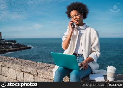 Portrait of young latin woman using her laptop and talking on the phone while sitting against the sea. Technology concept.