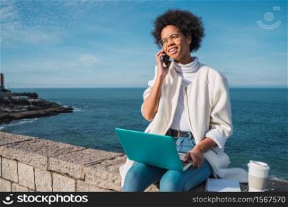 Portrait of young latin woman using her laptop and talking on the phone while sitting against the sea. Technology concept.