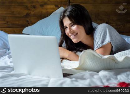 Portrait of young latin woman using her laptop while lying on the bed at home. Lifestyle concept