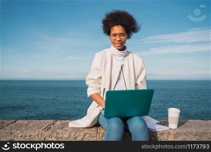 Portrait of young latin woman using her laptop while sitting against the sea. Technology concept.