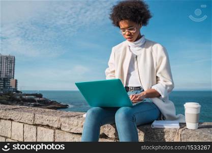 Portrait of young latin woman using her laptop while sitting against the sea. Technology concept.