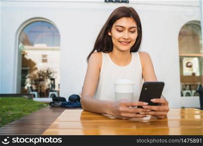 Portrait of young latin woman using her mobile phone while sitting at coffee shop outdoors. Communication concept.