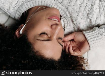 Portrait of young latin woman wearing earphones while lying in bed at home. Girl relaxing on bed.