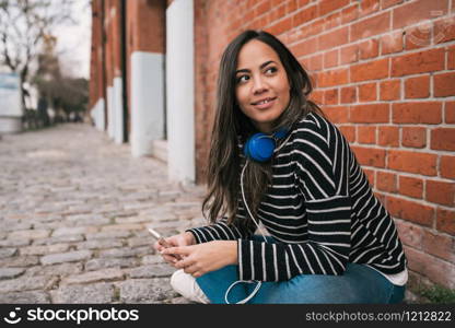 Portrait of young latin woman with headphones and using her mobile phone in the street. Outdoors.