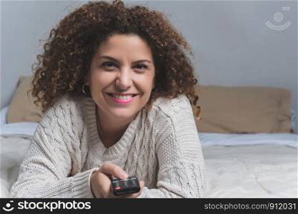 Portrait of young latin woman with remote lying in bed and watching tv. Television concept