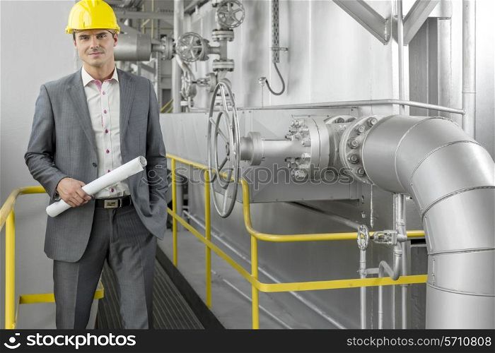 Portrait of young male architect holding blueprint by machinery at industry