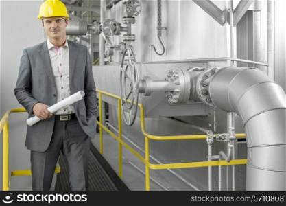 Portrait of young male architect holding blueprint by machinery at industry