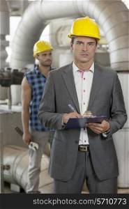 Portrait of young male manager writing on clipboard with manual worker in background at industry