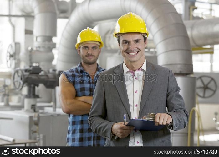 Portrait of young male supervisor holding clipboard with manual worker in background at industry