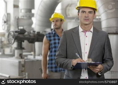 Portrait of young male supervisor writing on clipboard with manual worker in background at industry
