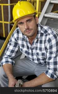 Portrait of young male worker in hard hat sitting on steps at industry