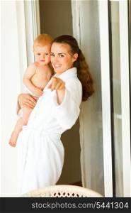 Portrait of young mama in bathrobe with baby in hand pointing in camera&#xA;
