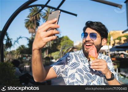 Portrait of young man enjoying sunny weather, taking a selfie with the phone and eating an ice cream outdoors.