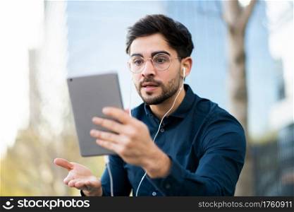 Portrait of young man having a video call on digital tablet while standing on bench outdoors. Urban concept.