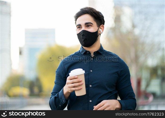 Portrait of young man holding a cup of coffee while standing outdoors on the street. New normal lifestyle concept. 