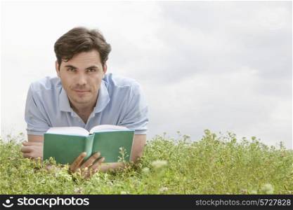 Portrait of young man holding book while lying on grass against sky