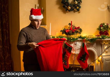 Portrait of young man in red hat looking inside of Santa bag with amazement