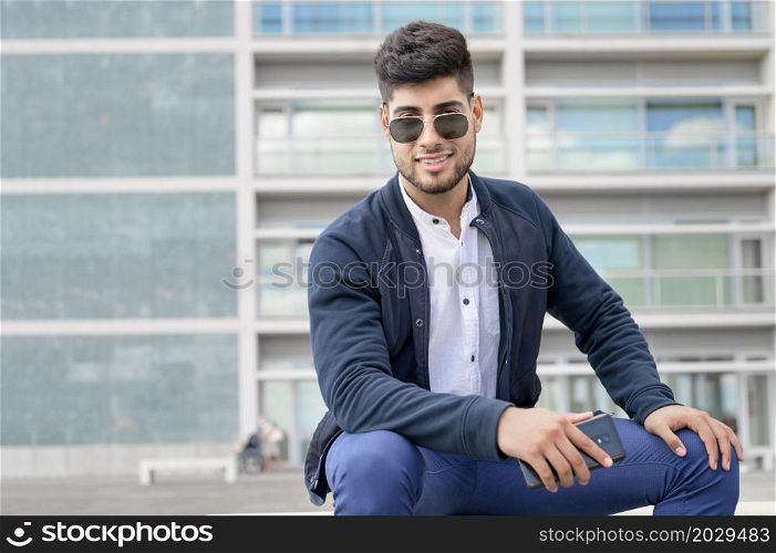 Portrait of young man in stylish sunglasses posing while sitting in bench in urban background. High quality photo. Portrait of young man in stylish sunglasses posing while sitting in bench in urban background.