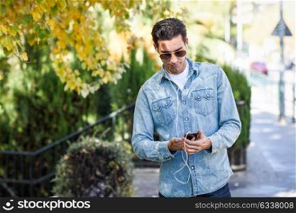 Portrait of young man in urban background listening to music with headphones. Guy wearing casual clothes and aviator sunglasses with a smartphone.