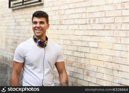 Portrait of young man in urban background smiling with headphones. Wearing white t-shirt near a brick wall