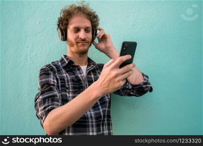 Portrait of young man listening to music with headphones and mobile phone against light blue background.