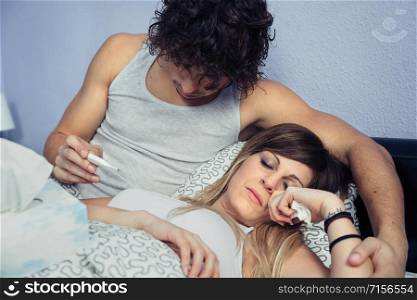 Portrait of young man looking temperature in thermometer of sick woman lying on bed. Sickness and healthcare concept.. Man looking temperature of sick woman in thermometer