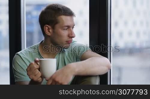 Portrait of young man sitting on the window with coffee cup, looking at dawn city scenery after early wakeup. Handsome casual guy relaxing in penthouse after good night sleep and watching sunrise.