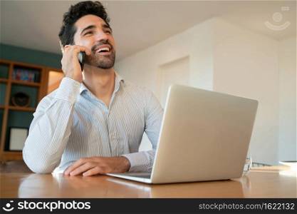 Portrait of young man talking on his mobile phone and working from home with laptop. Home office concept. New normal lifestyle.
