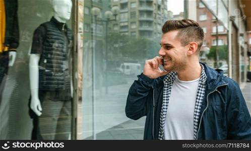 Portrait of young man talking on the cell phone looking at the showcase of a fashion store. Man looking at the showcase of a store