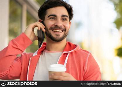 Portrait of young man talking on the phone and drinking coffee while walking outdoors on the street. Urban concept.