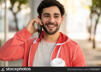 Portrait of young man talking on the phone and drinking coffee while walking outdoors on the street. Urban concept.