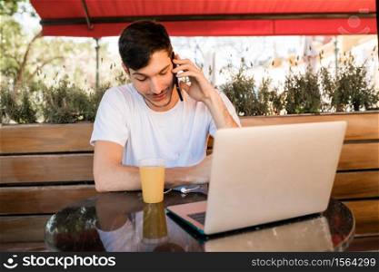 Portrait of young man talking on the phone and using laptp while sitting in a coffee shop outdoors. Communication concept.