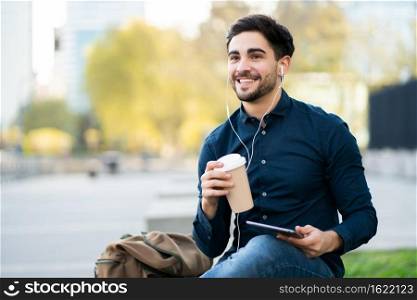 Portrait of young man using digital tablet and holding a cup of coffee while sitting on bench outdoors. Urban concept.