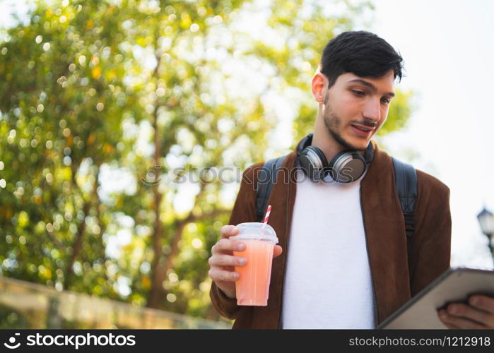 Portrait of young man using his digital tablet while drinking fresh fruit juice outdoors in the street. Technology and urban concept.
