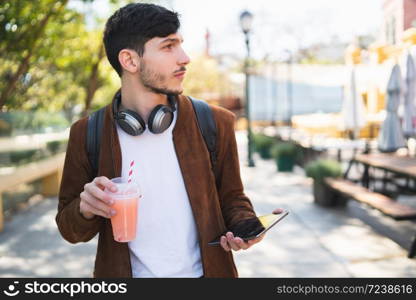 Portrait of young man using his digital tablet while drinking fresh fruit juice outdoors in the street. Technology and urban concept.