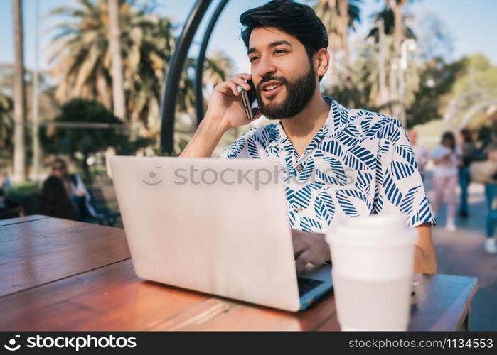 Portrait of young man using his laptop and talking on the phone while sitting in a coffee shop. Technology and business concept.