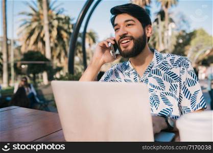 Portrait of young man using his laptop and talking on the phone while sitting in a coffee shop. Technology and business concept.