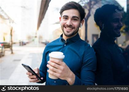 Portrait of young man using his mobile phone and holding a cup of coffee while standing outdoors at the street. Urban concept.