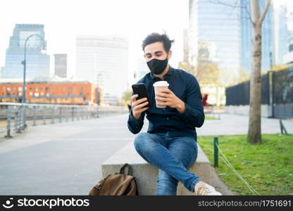 Portrait of young man using his mobile phone and holding a cup of coffee while sitting on bench outdoors. New normal lifestyle concept. Urban concept.