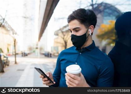 Portrait of young man using his mobile phone and holding a cup of coffee while standing outdoors at the street. New normal lifestyle concept. Urban concept.