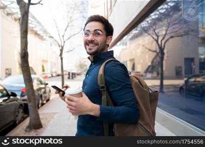 Portrait of young man using his mobile phone and holding a cup of coffee while walking outdoors at the street. Urban concept.