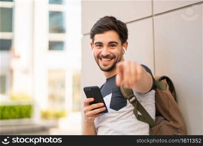 Portrait of young man using his mobile phone and pointing to camera outdoors. Urban concept.. Young man using his mobile phone outdoors.