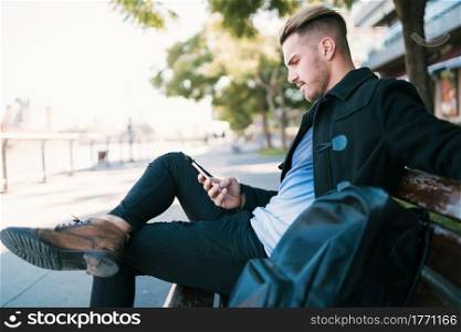 Portrait of young man using his mobile phone outdoors while sitting on a bench. Communication concept.